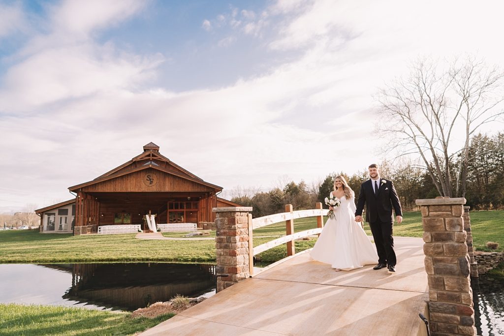 Newlyweds walking over bridge with The Barn at Sycamore Farms behind them at Sunset