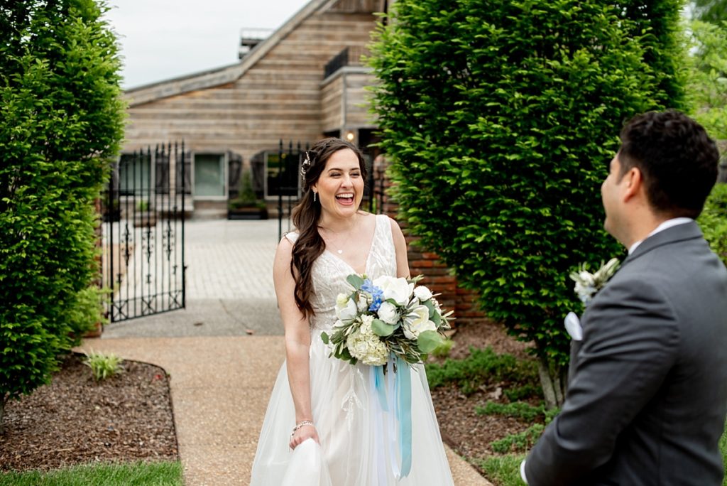 Bride smiling at her fiance during their first look, she is holding a white and blue bouquet