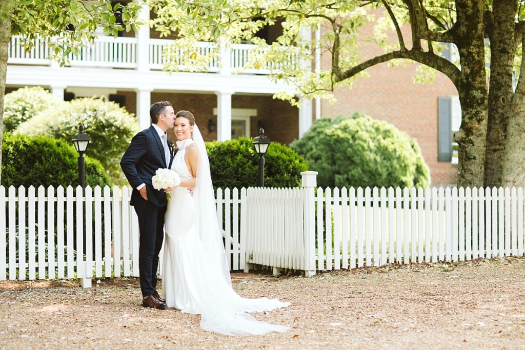 Groom kissing his bride on the forehead by a white picket fence